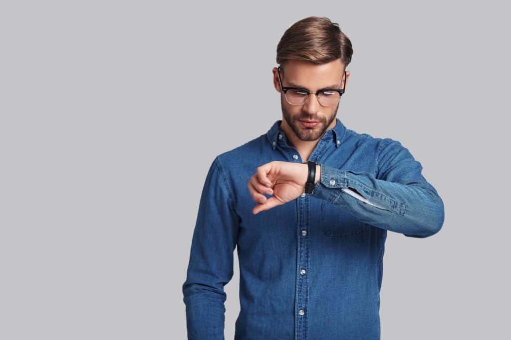 young man in eyewear checking the time while standing against grey background